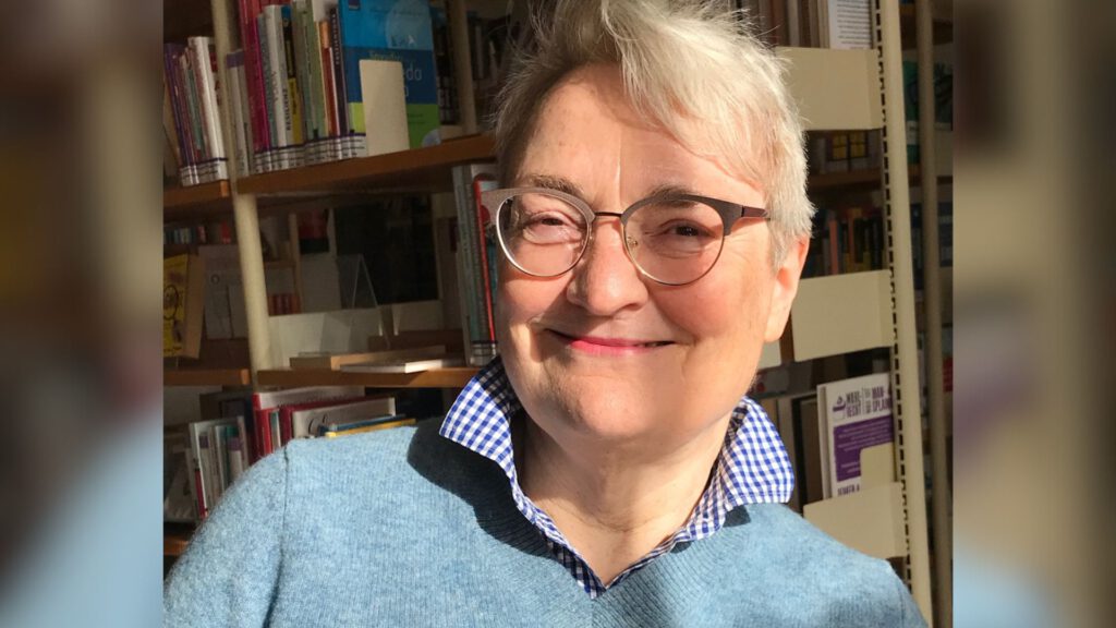 Eine Frau mit kurzen grauen Haaren und Brille lächelt vor einem Bücherregal in einer Bibliothek.