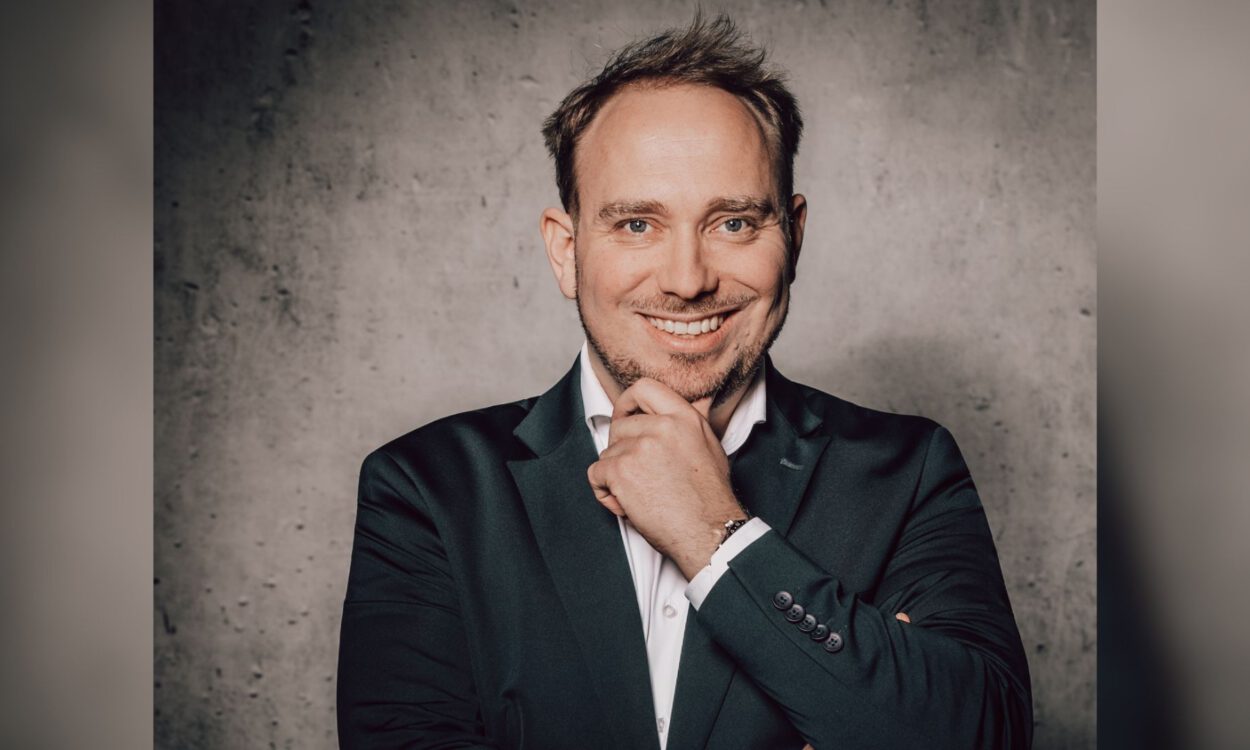 Ein Mann im Anzug lächelt und stützt nachdenklich das Kinn auf seine Hand vor einer grauen Wand.