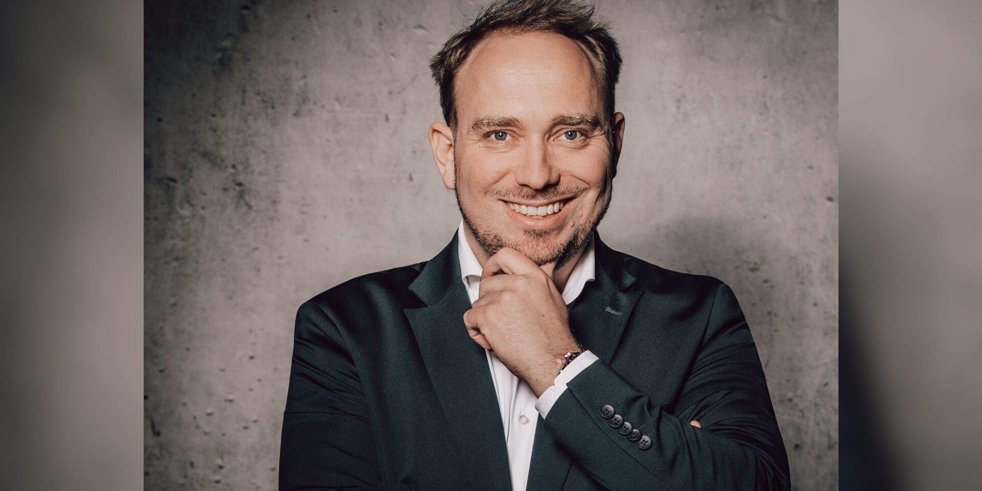 Ein Mann im Anzug lächelt und stützt nachdenklich das Kinn auf seine Hand vor einer grauen Wand.