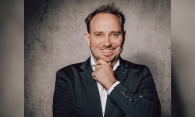 Ein Mann im Anzug lächelt und stützt nachdenklich das Kinn auf seine Hand vor einer grauen Wand.