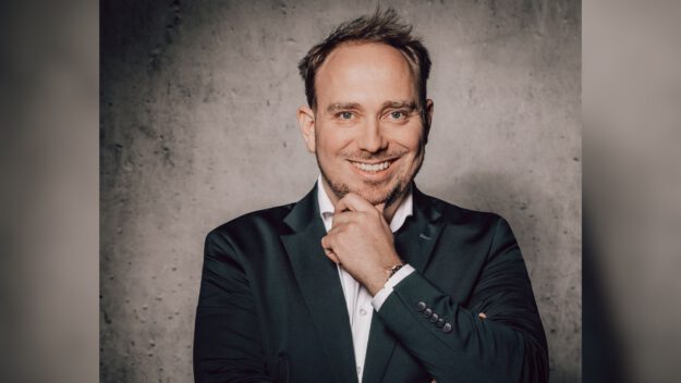 Ein Mann im Anzug lächelt und stützt nachdenklich das Kinn auf seine Hand vor einer grauen Wand.