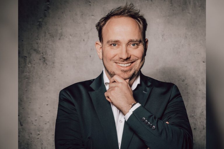 Ein Mann im Anzug lächelt und stützt nachdenklich das Kinn auf seine Hand vor einer grauen Wand.