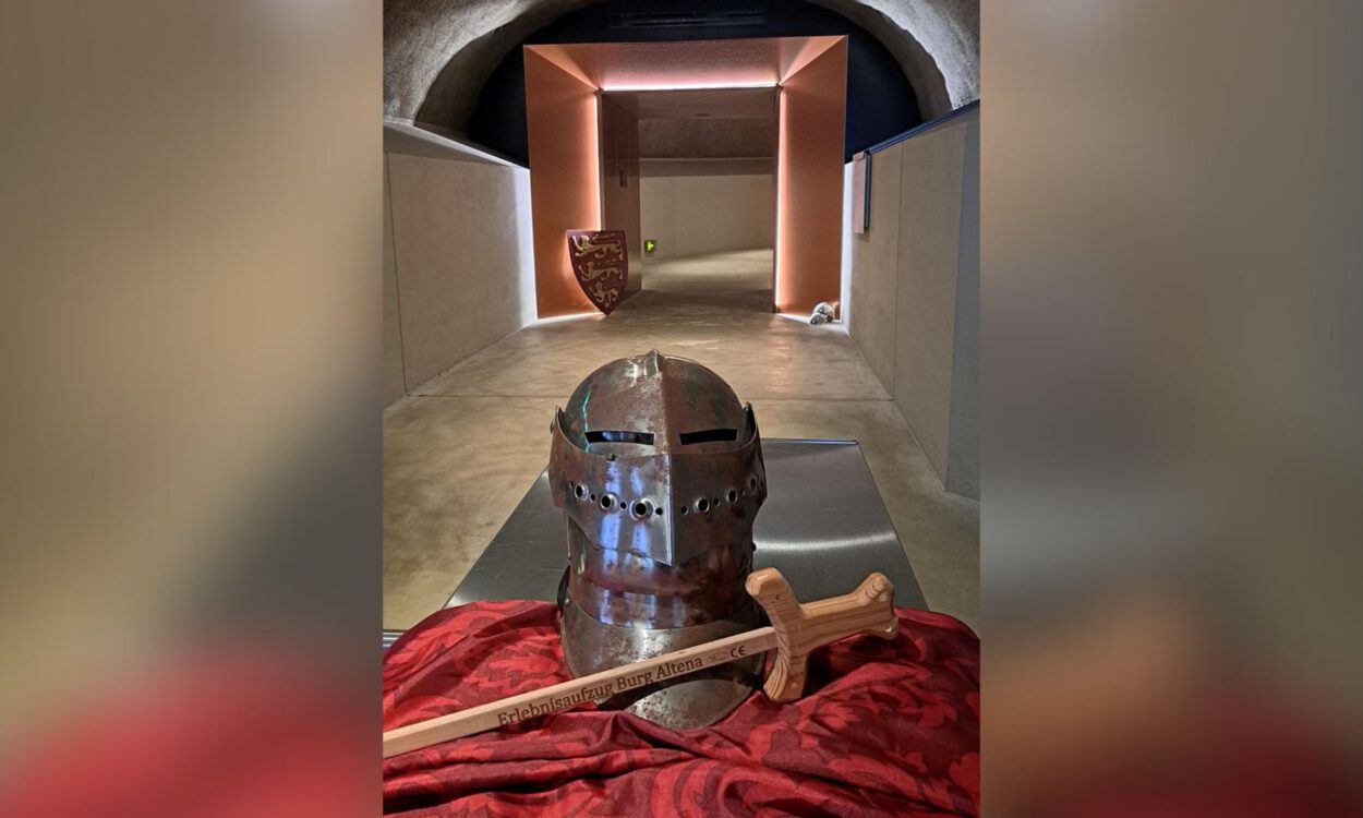 Ein Ritterhelm und ein Holzschwert liegen auf einem Tuch in einem gewölbten Gang mit beleuchtetem Durchgang.
