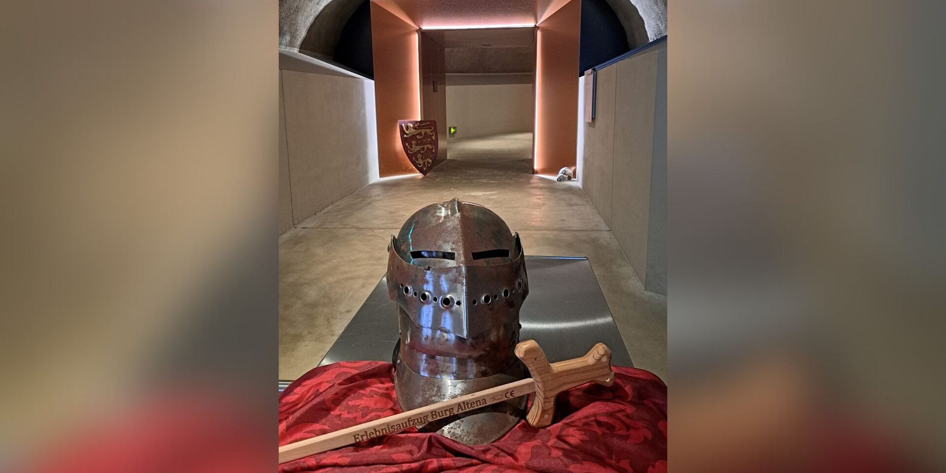 Ein Ritterhelm und ein Holzschwert liegen auf einem Tuch in einem gewölbten Gang mit beleuchtetem Durchgang.