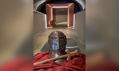 Ein Ritterhelm und ein Holzschwert liegen auf einem Tuch in einem gewölbten Gang mit beleuchtetem Durchgang.