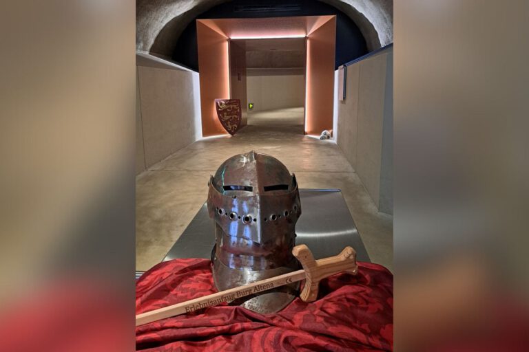 Ein Ritterhelm und ein Holzschwert liegen auf einem Tuch in einem gewölbten Gang mit beleuchtetem Durchgang.