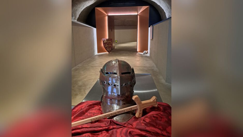 Ein Ritterhelm und ein Holzschwert liegen auf einem Tuch in einem gewölbten Gang mit beleuchtetem Durchgang.