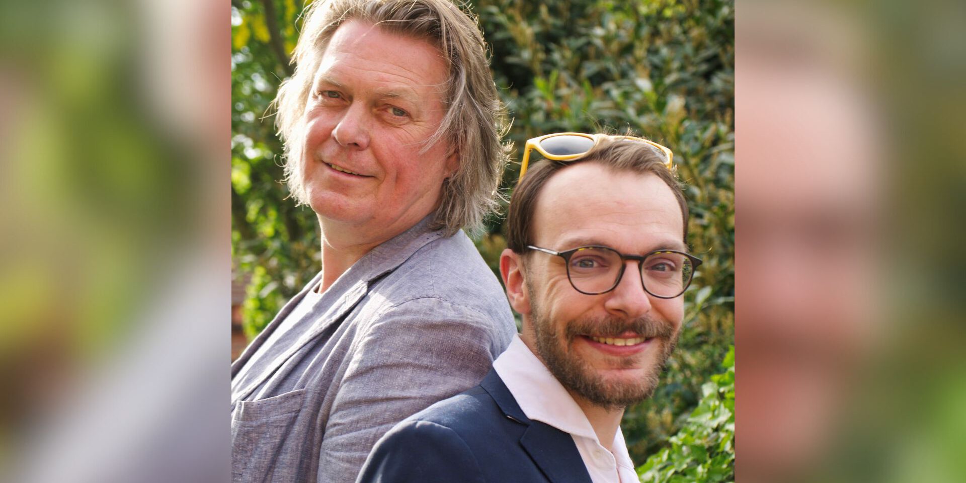 Zwei Männer stehen Rücken an Rücken im Freien und lächeln in die Kamera, umgeben von grüner Vegetation.