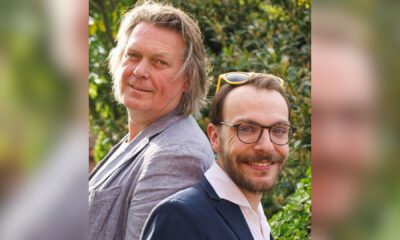 Zwei Männer stehen Rücken an Rücken im Freien und lächeln in die Kamera, umgeben von grüner Vegetation.