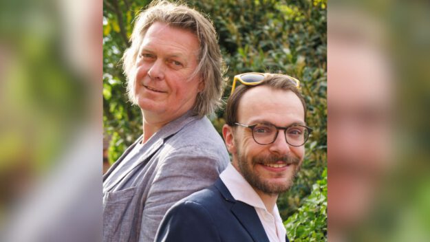 Zwei Männer stehen Rücken an Rücken im Freien und lächeln in die Kamera, umgeben von grüner Vegetation.