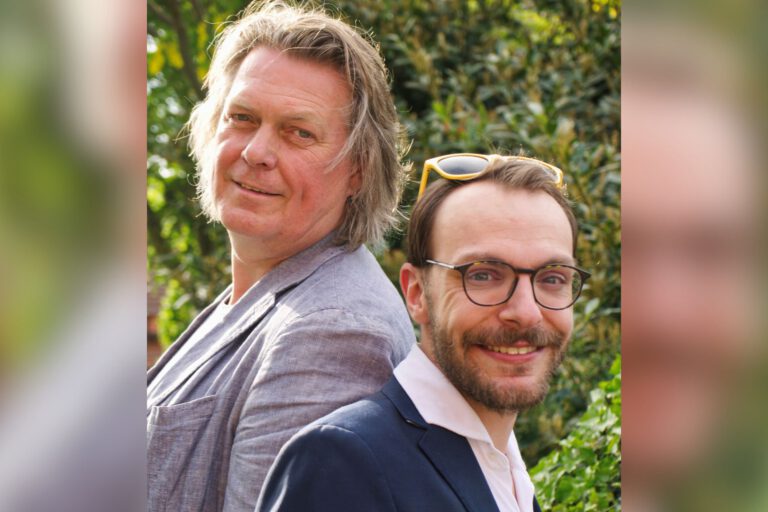 Zwei Männer stehen Rücken an Rücken im Freien und lächeln in die Kamera, umgeben von grüner Vegetation.