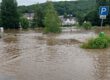 Überflutete Straße mit braunem Hochwasser, ein Parkplatzschild ragt aus dem Wasser, im Hintergrund Häuser und Bäume teilweise von der Flut umgeben.