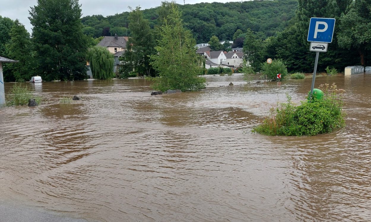 Überflutete Straße mit braunem Hochwasser, ein Parkplatzschild ragt aus dem Wasser, im Hintergrund Häuser und Bäume teilweise von der Flut umgeben.