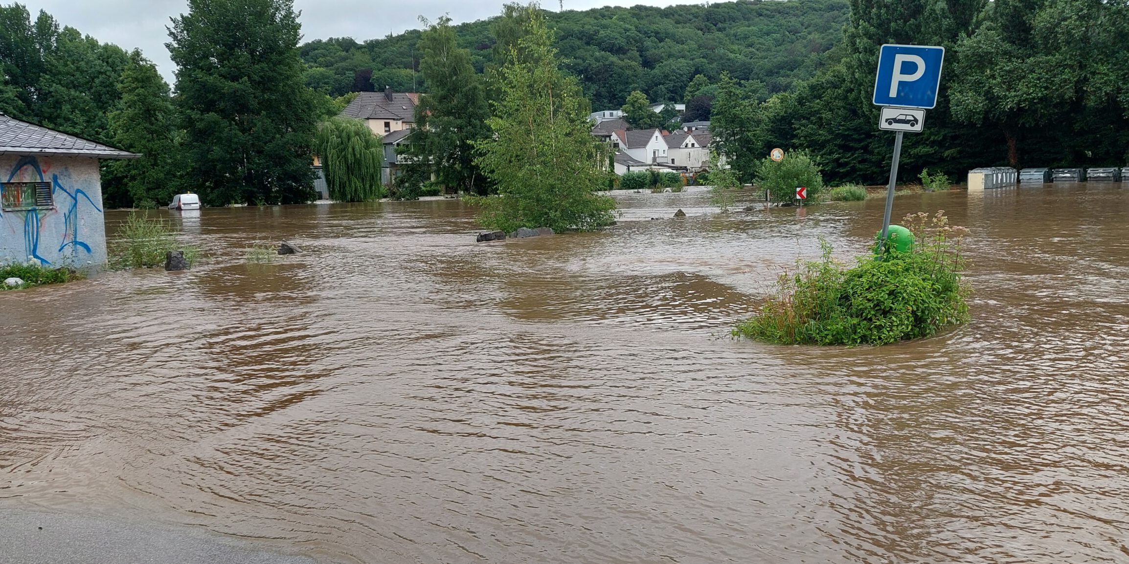 Überflutete Straße mit braunem Hochwasser, ein Parkplatzschild ragt aus dem Wasser, im Hintergrund Häuser und Bäume teilweise von der Flut umgeben.