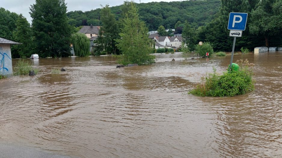 Überflutete Straße mit braunem Hochwasser, ein Parkplatzschild ragt aus dem Wasser, im Hintergrund Häuser und Bäume teilweise von der Flut umgeben.
