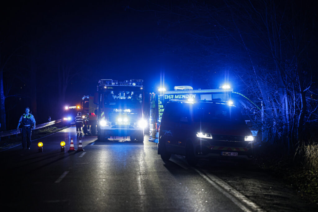 Feuerwehr- und Rettungsfahrzeuge mit Blaulicht stehen nachts auf einer gesperrten Landstraße; Einsatzkräfte sichern die Unfallstelle ab.