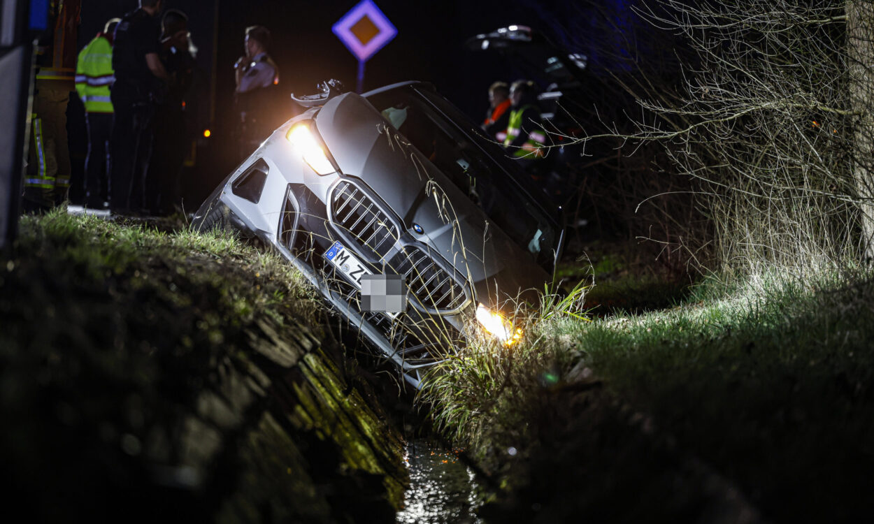 Auto ist nachts von der Straße abgekommen und in einen Graben gerutscht; Einsatzkräfte stehen im Hintergrund, die Scheinwerfer leuchten.
