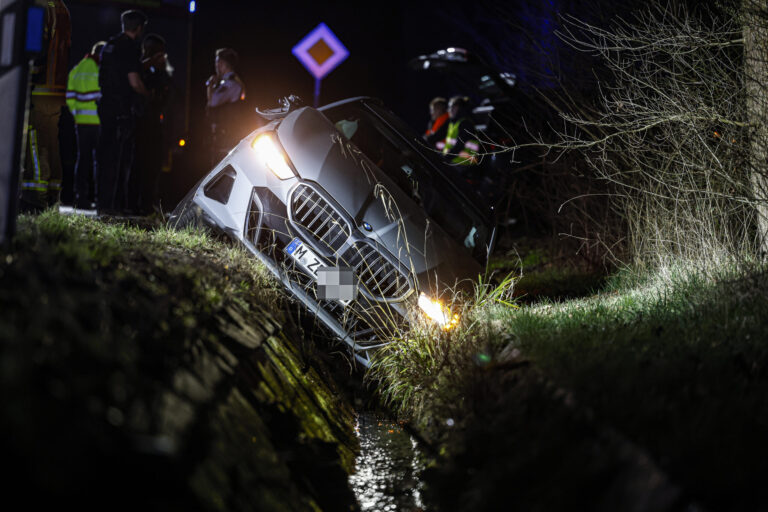 Auto ist nachts von der Straße abgekommen und in einen Graben gerutscht; Einsatzkräfte stehen im Hintergrund, die Scheinwerfer leuchten.