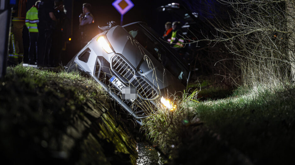 Auto ist nachts von der Straße abgekommen und in einen Graben gerutscht; Einsatzkräfte stehen im Hintergrund, die Scheinwerfer leuchten.
