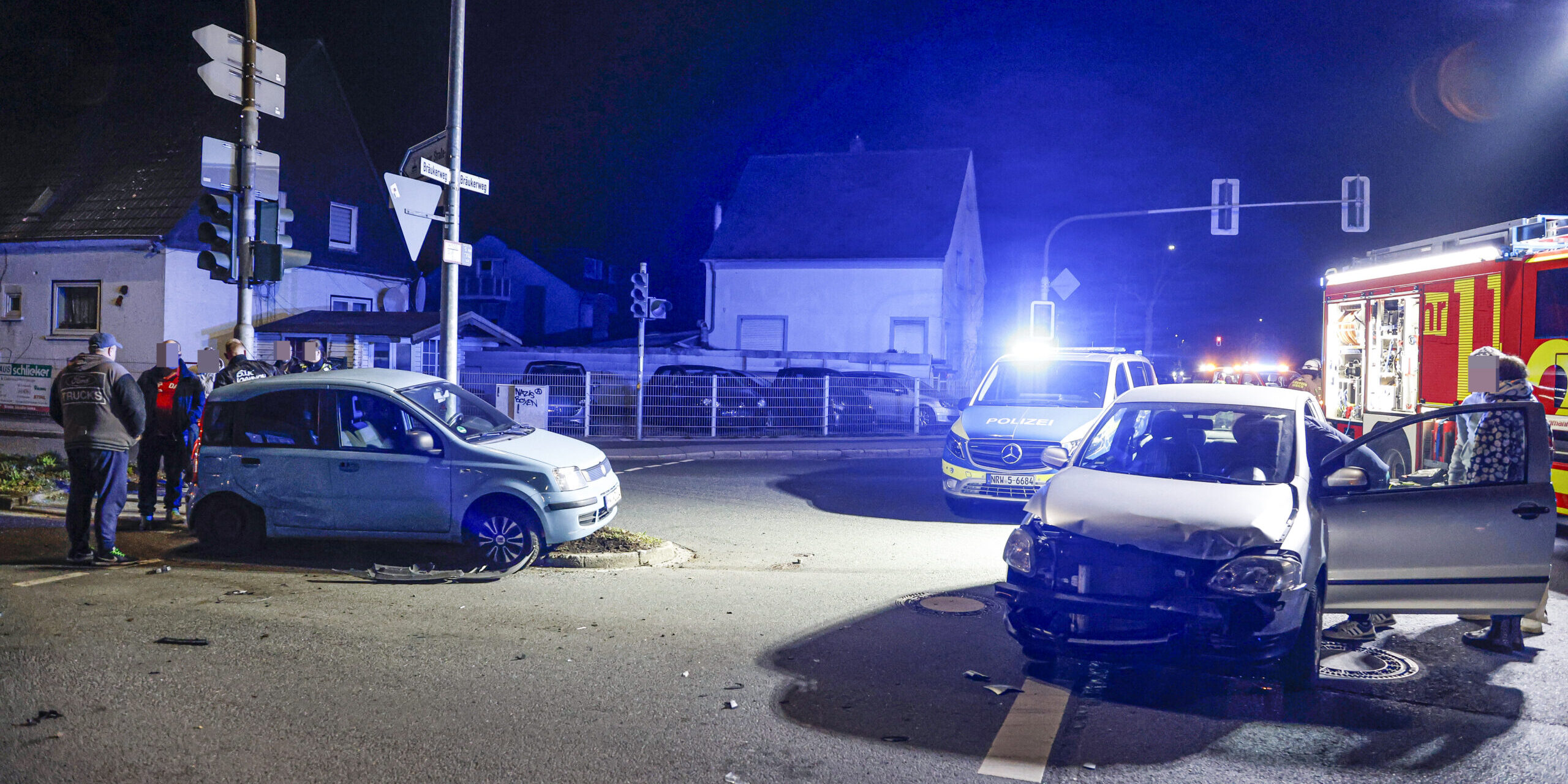 Zwei beschädigte Autos stehen nachts nach einem Unfall auf einer Kreuzung; Polizei und Feuerwehr sind mit Blaulicht vor Ort.