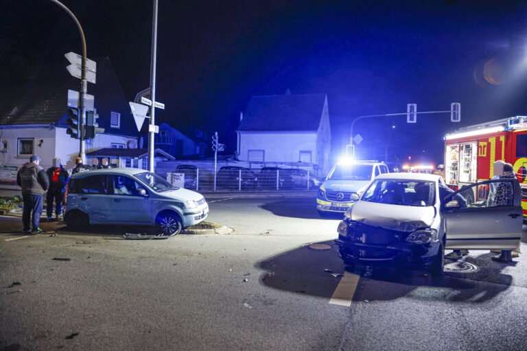 Zwei beschädigte Autos stehen nachts nach einem Unfall auf einer Kreuzung; Polizei und Feuerwehr sind mit Blaulicht vor Ort.