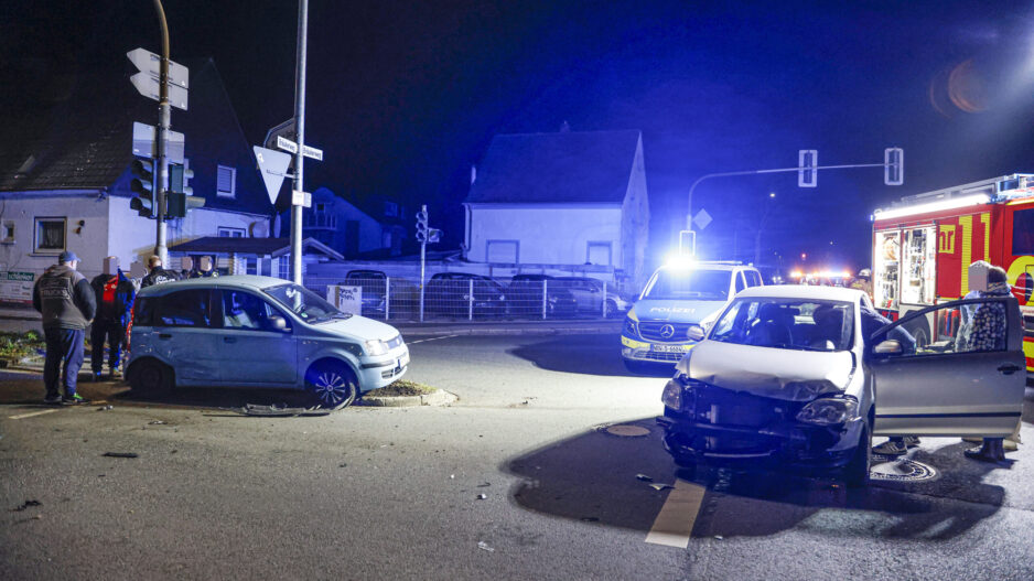 Zwei beschädigte Autos stehen nachts nach einem Unfall auf einer Kreuzung; Polizei und Feuerwehr sind mit Blaulicht vor Ort.