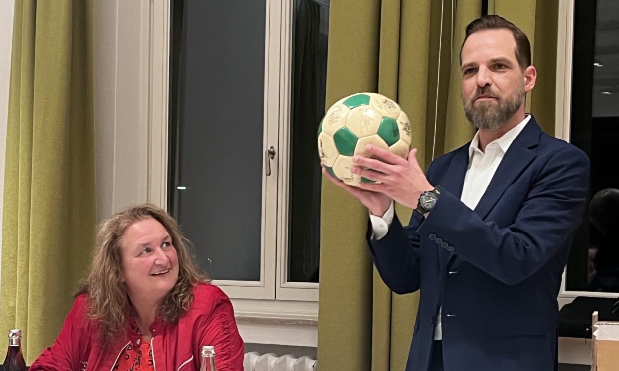 Ein Mann im Anzug hält bei einer Sitzung einen grün-weißen Ball hoch, während eine Frau am Tisch ihm aufmerksam zusieht.