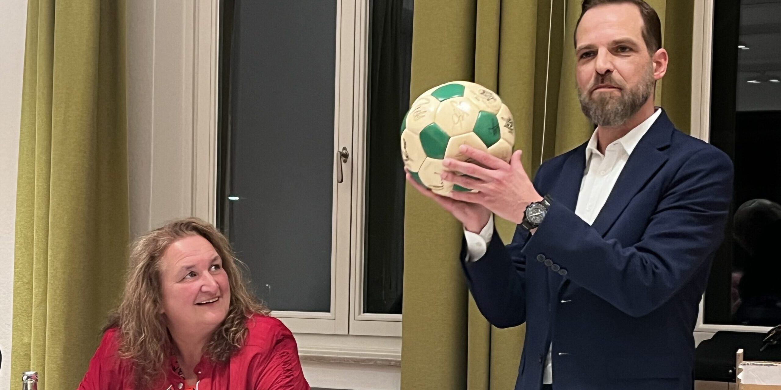 Ein Mann im Anzug hält bei einer Sitzung einen grün-weißen Ball hoch, während eine Frau am Tisch ihm aufmerksam zusieht.