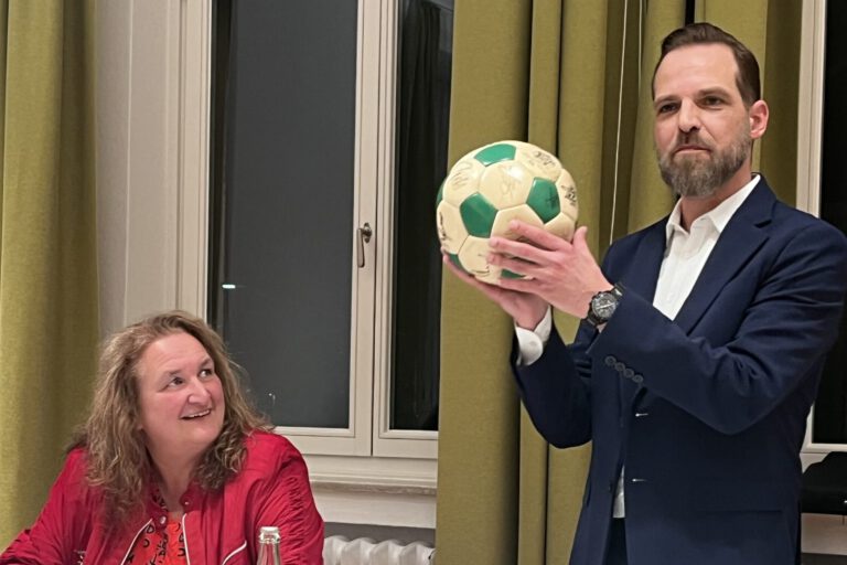 Ein Mann im Anzug hält bei einer Sitzung einen grün-weißen Ball hoch, während eine Frau am Tisch ihm aufmerksam zusieht.