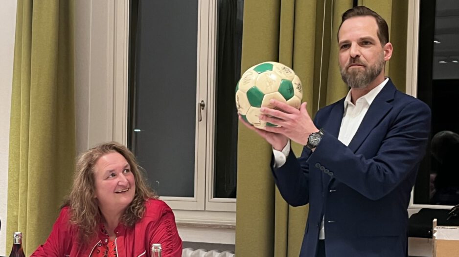 Ein Mann im Anzug hält bei einer Sitzung einen grün-weißen Ball hoch, während eine Frau am Tisch ihm aufmerksam zusieht.