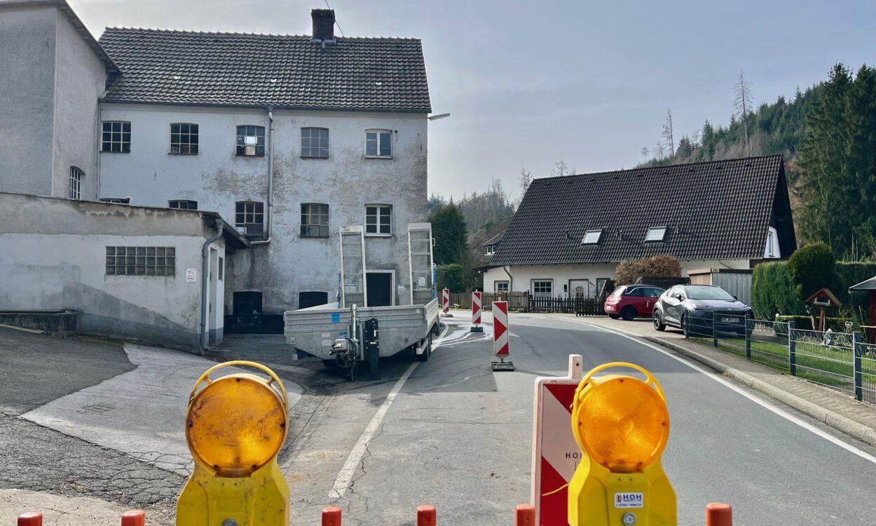 Eine teilweise abgesperrte Straße führt an einem alten Gebäude vorbei; Warnbaken und Baustellenleuchten stehen auf der Fahrbahn.
