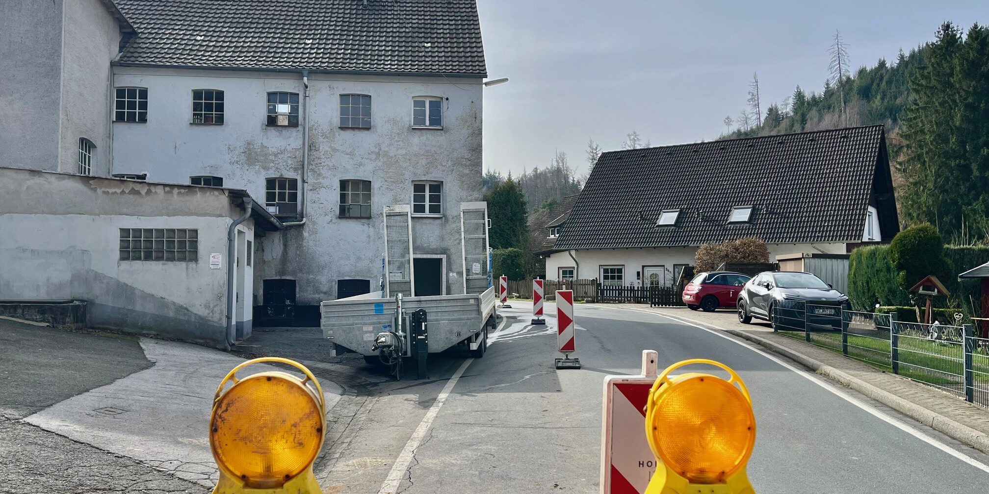 Eine teilweise abgesperrte Straße führt an einem alten Gebäude vorbei; Warnbaken und Baustellenleuchten stehen auf der Fahrbahn.