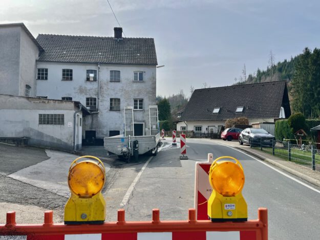 Eine teilweise abgesperrte Straße führt an einem alten Gebäude vorbei; Warnbaken und Baustellenleuchten stehen auf der Fahrbahn.