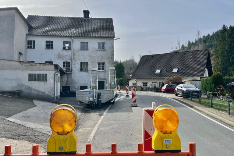 Eine teilweise abgesperrte Straße führt an einem alten Gebäude vorbei; Warnbaken und Baustellenleuchten stehen auf der Fahrbahn.