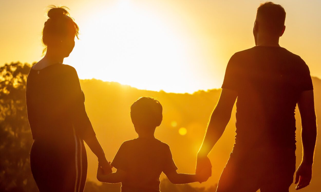 Zwei Erwachsene und ein Kind halten sich bei Sonnenuntergang an den Händen und schauen in die Ferne.