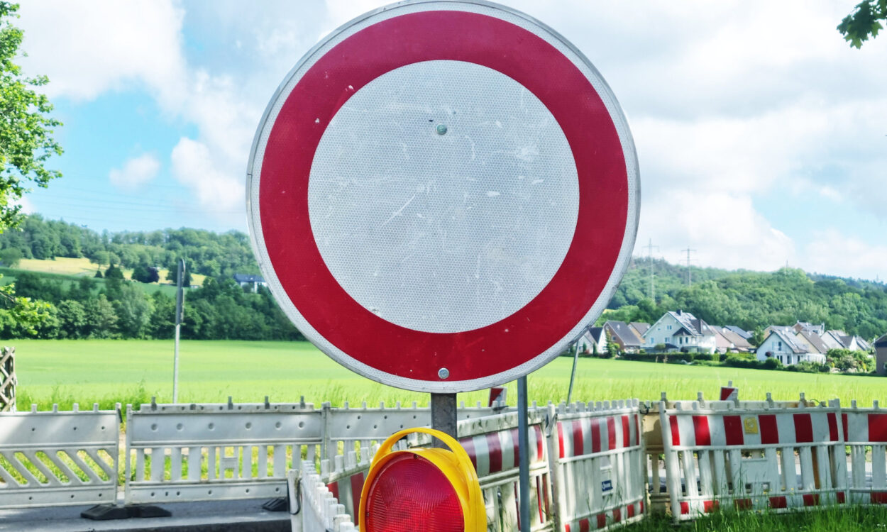 Ein rundes Verkehrsschild „Durchfahrt verboten“ steht vor einer abgesperrten Straße mit Baustellenbarrieren in ländlicher Umgebung.