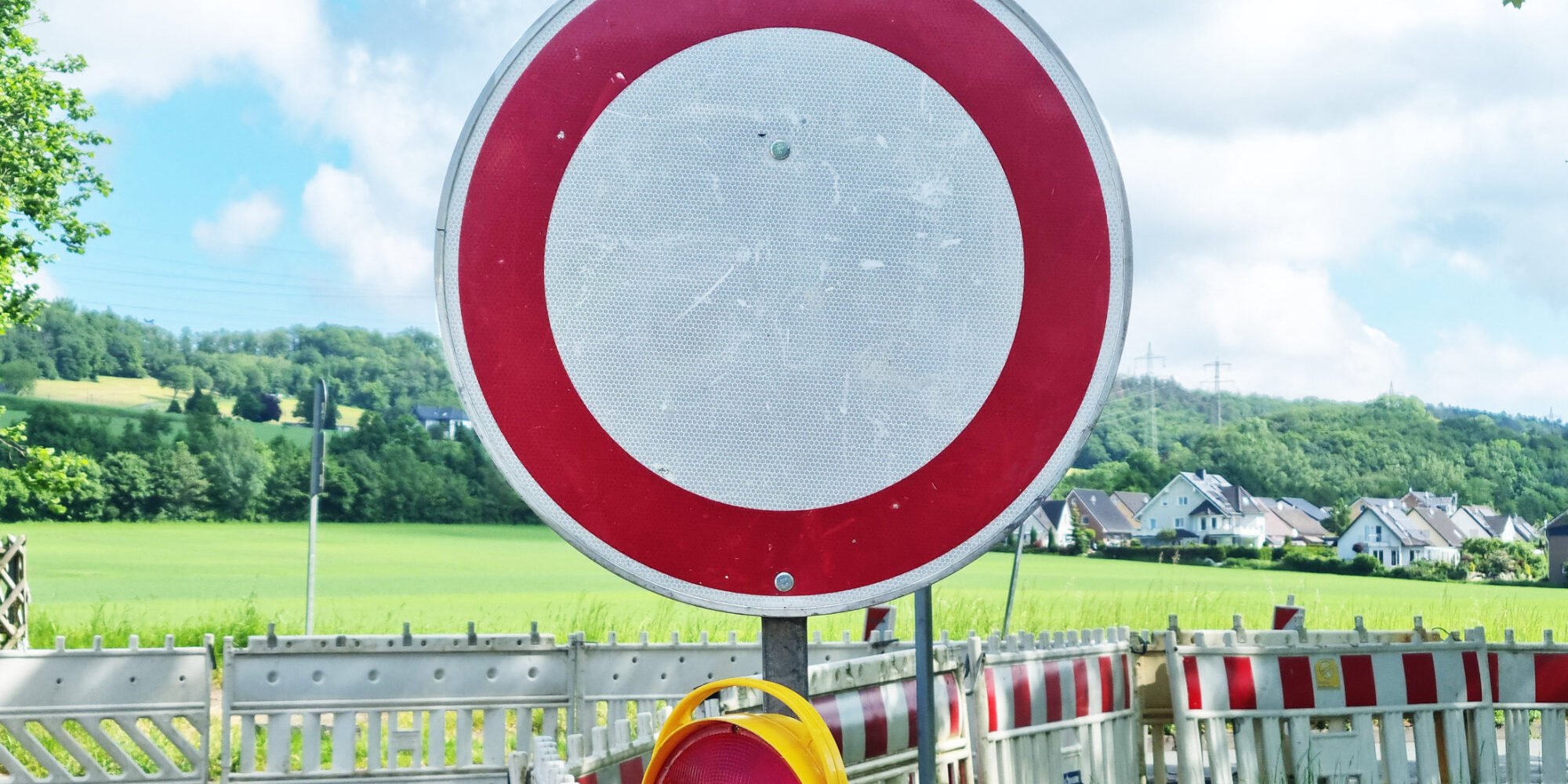 Ein rundes Verkehrsschild „Durchfahrt verboten“ steht vor einer abgesperrten Straße mit Baustellenbarrieren in ländlicher Umgebung.