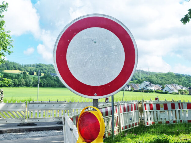 Ein rundes Verkehrsschild „Durchfahrt verboten“ steht vor einer abgesperrten Straße mit Baustellenbarrieren in ländlicher Umgebung.