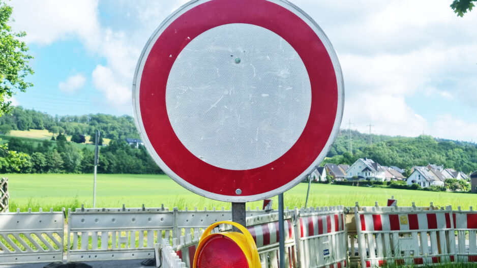 Ein rundes Verkehrsschild „Durchfahrt verboten“ steht vor einer abgesperrten Straße mit Baustellenbarrieren in ländlicher Umgebung.