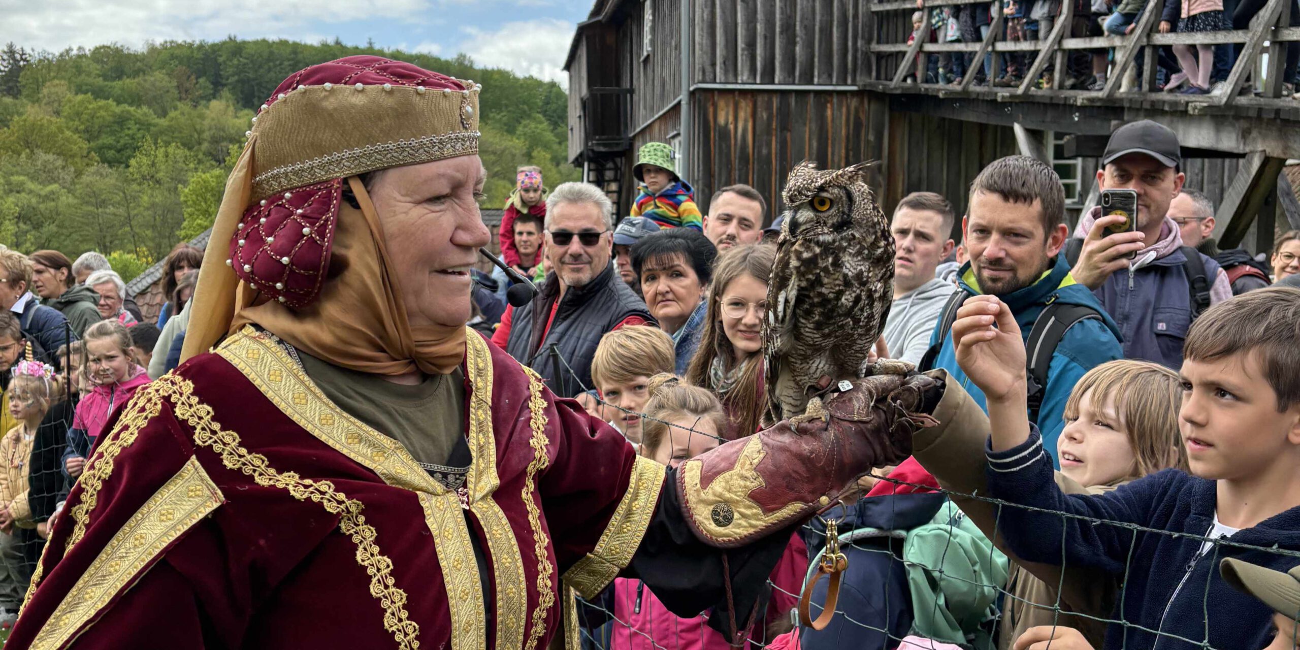 Frau in historischer Kleidung präsentiert eine Eule auf ihrer Hand vor einer Gruppe von Kindern und Erwachsenen bei einer Vorführung im Freien.