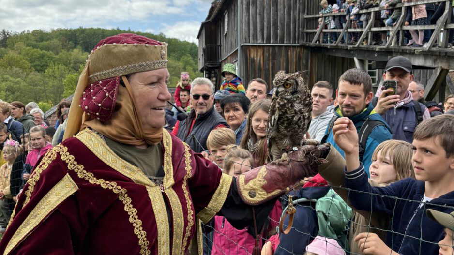 Frau in historischer Kleidung präsentiert eine Eule auf ihrer Hand vor einer Gruppe von Kindern und Erwachsenen bei einer Vorführung im Freien.
