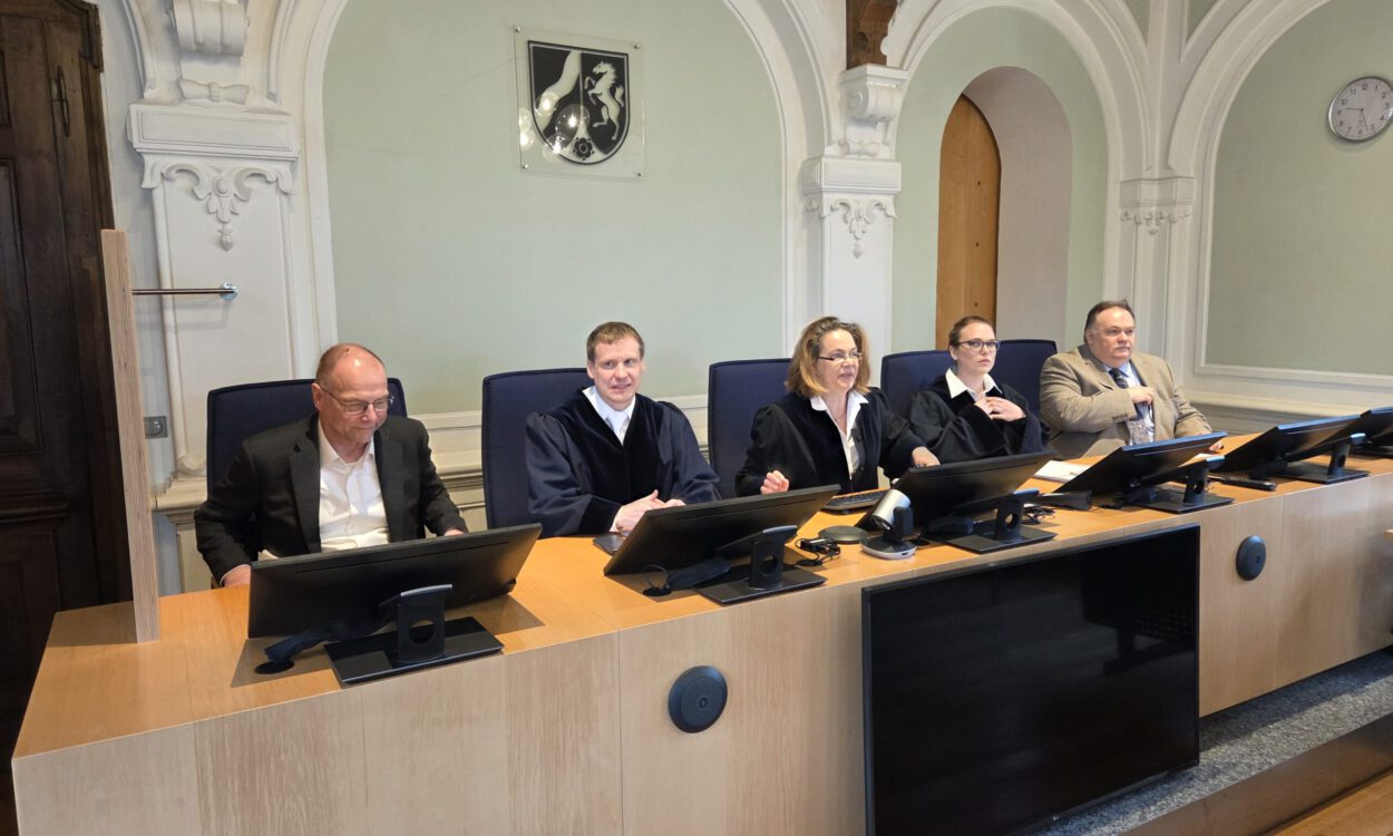 Mehrere Personen sitzen an einem Richtertisch in einem Gerichtssaal; drei tragen Richterroben, im Hintergrund sind hohe Rundbögen und das Landeswappen zu sehen.