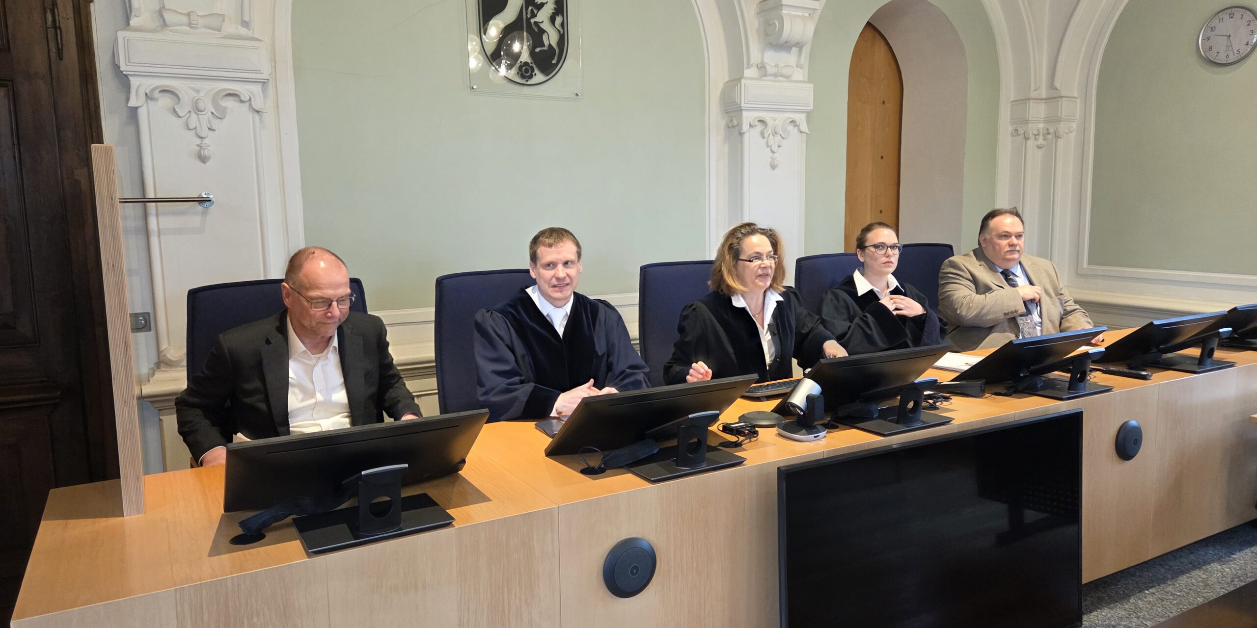 Mehrere Personen sitzen an einem Richtertisch in einem Gerichtssaal; drei tragen Richterroben, im Hintergrund sind hohe Rundbögen und das Landeswappen zu sehen.