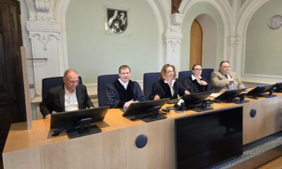 Mehrere Personen sitzen an einem Richtertisch in einem Gerichtssaal; drei tragen Richterroben, im Hintergrund sind hohe Rundbögen und das Landeswappen zu sehen.