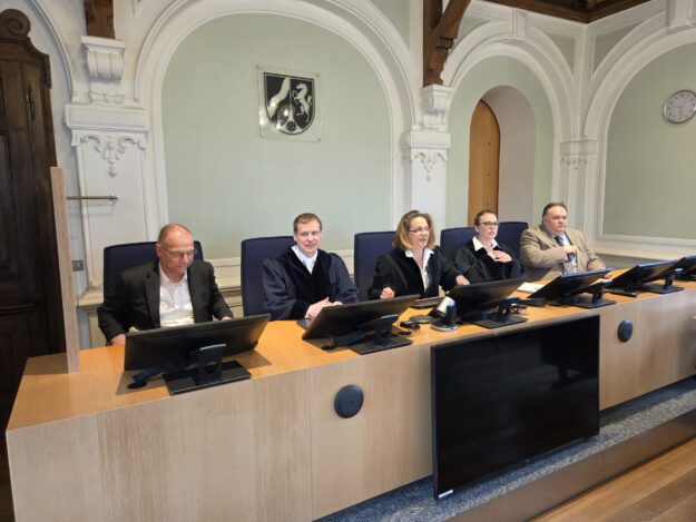 Mehrere Personen sitzen an einem Richtertisch in einem Gerichtssaal; drei tragen Richterroben, im Hintergrund sind hohe Rundbögen und das Landeswappen zu sehen.