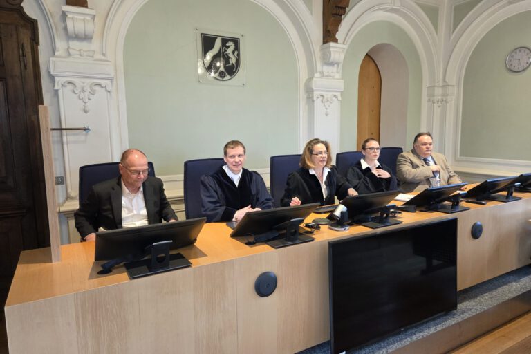 Mehrere Personen sitzen an einem Richtertisch in einem Gerichtssaal; drei tragen Richterroben, im Hintergrund sind hohe Rundbögen und das Landeswappen zu sehen.