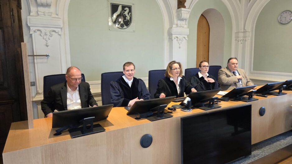 Mehrere Personen sitzen an einem Richtertisch in einem Gerichtssaal; drei tragen Richterroben, im Hintergrund sind hohe Rundbögen und das Landeswappen zu sehen.