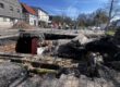 Großes Loch in einer Straße mit freigelegten Rohren und abgesacktem Asphalt; die Baustelle ist abgesperrt, im Hintergrund stehen Bagger und Gebäude.