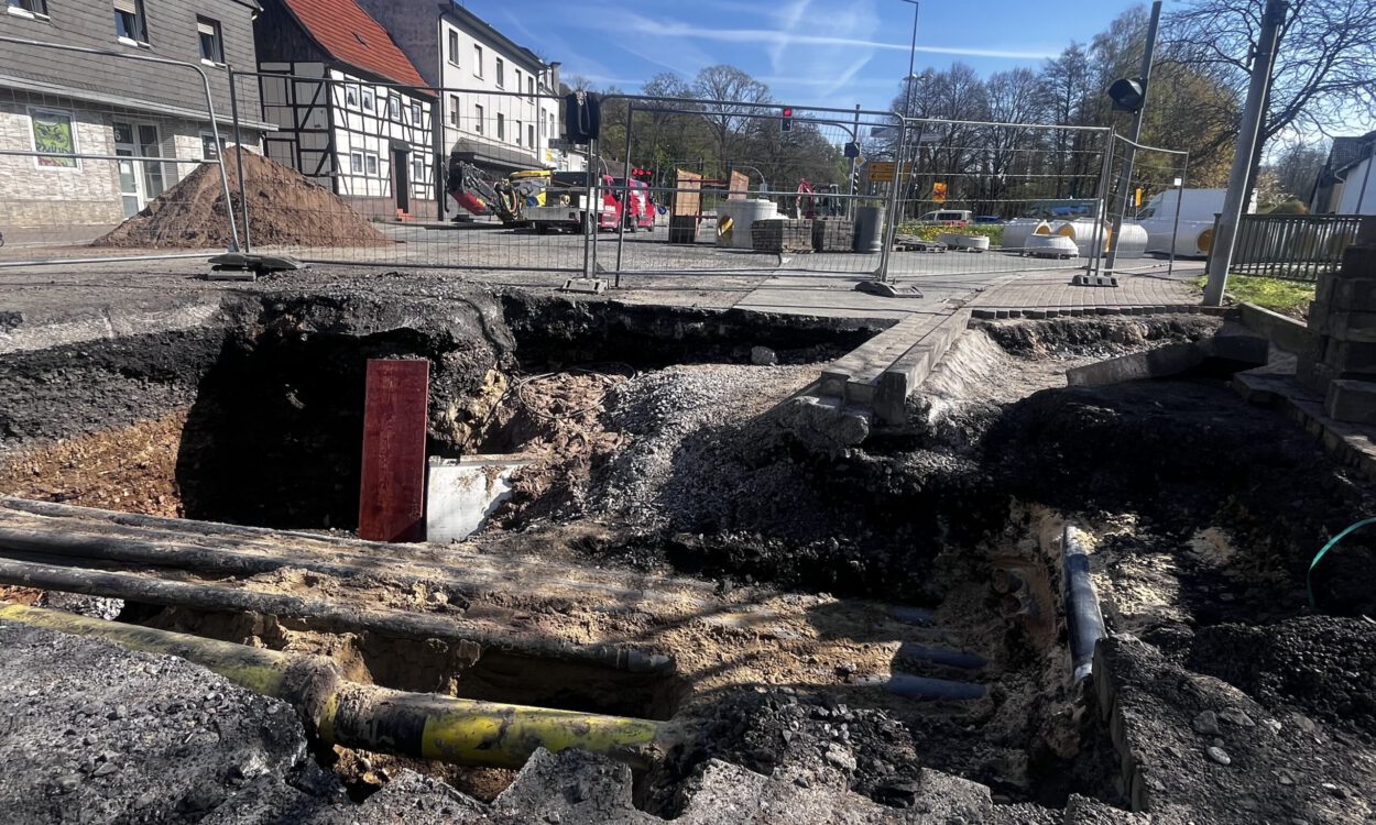 Großes Loch in einer Straße mit freigelegten Rohren und abgesacktem Asphalt; die Baustelle ist abgesperrt, im Hintergrund stehen Bagger und Gebäude.