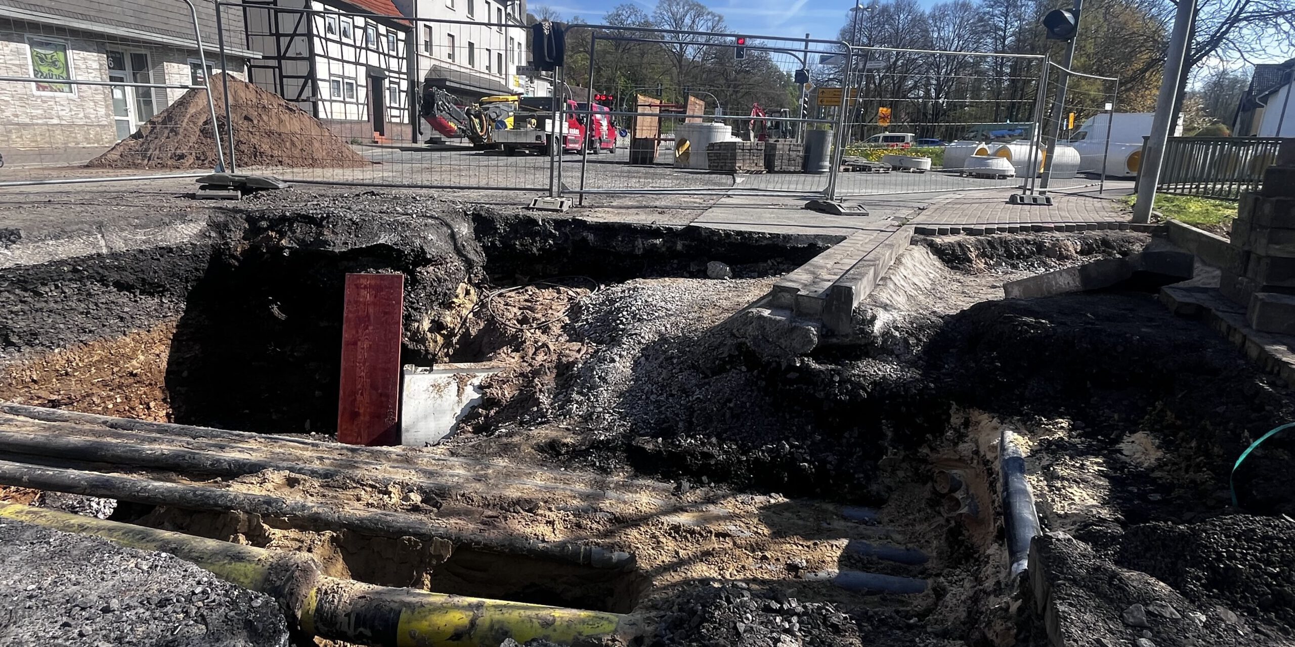 Großes Loch in einer Straße mit freigelegten Rohren und abgesacktem Asphalt; die Baustelle ist abgesperrt, im Hintergrund stehen Bagger und Gebäude.